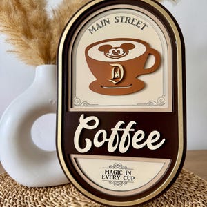 May include: A brown oval-shaped wooden sign with gold trim. The sign reads "Main Street Coffee" with a Mickey Mouse head in a coffee cup. The text "Magic in Every Cup" is below the coffee cup.