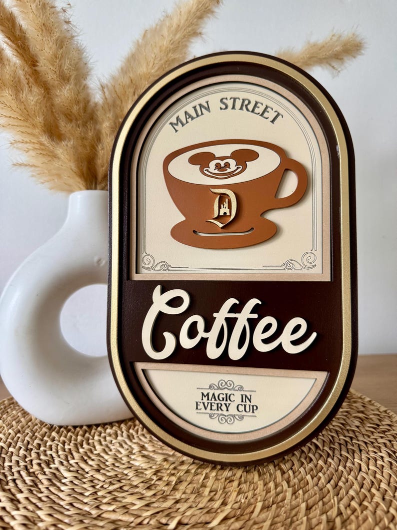 May include: A brown oval-shaped wooden sign with gold trim. The sign reads "Main Street Coffee" with a Mickey Mouse head in a coffee cup. The text "Magic in Every Cup" is below the coffee cup.