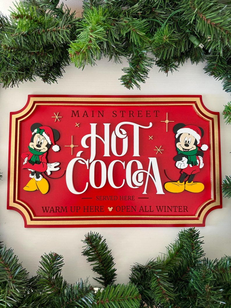 K&ouml;nnte beinhalten: Rotes Holzschild mit goldener Umrandung, auf dem Mickey und Minnie Mouse in Weihnachtsmannm&uuml;tzen zu sehen sind. Das Schild lautet "Main Street Hot Cocoa Served Here Warm Up Here Open All Winter".