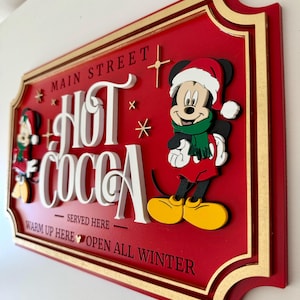 K&ouml;nnte beinhalten: Ein rotes Holzschild mit goldenem Rahmen und Mickey Mouse, der einen Weihnachtsmann-Hut tr&auml;gt. Das Schild lautet "Main Street Hot Cocoa - Served Here - Warm Up Here - Open All Winter".
