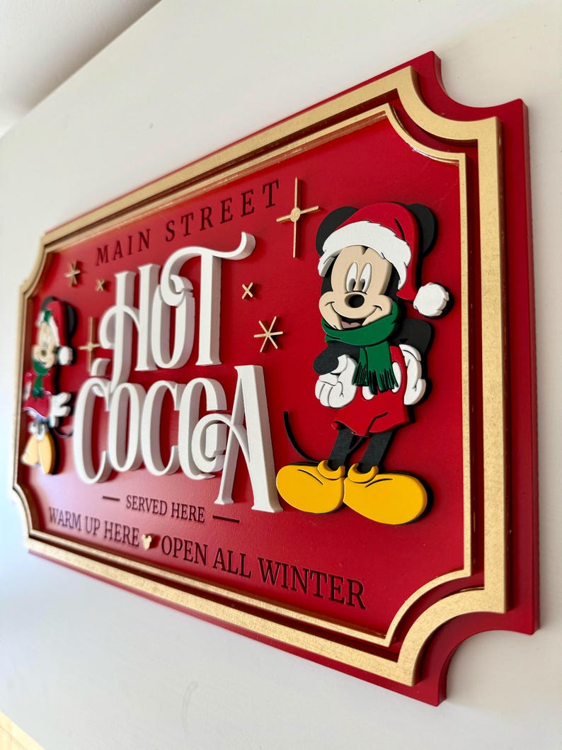 K&ouml;nnte beinhalten: Ein rotes Holzschild mit goldenem Rahmen und Mickey Mouse, der einen Weihnachtsmann-Hut tr&auml;gt. Das Schild lautet "Main Street Hot Cocoa - Served Here - Warm Up Here - Open All Winter".