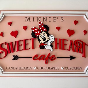 May include: A pink and white sign with a white border and a red and white Minnie Mouse illustration. The sign says "Minnie's Sweetheart cafe" with an arrow on either side of the word "cafe". The sign also says "Candy Hearts, Chocolates, Cupcakes" at the bottom.