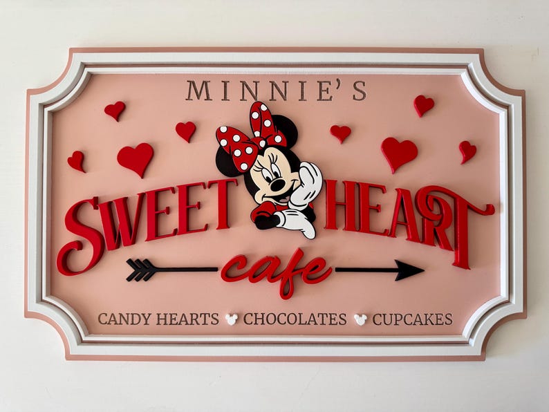 May include: A pink and white sign with a white border and a red and white Minnie Mouse illustration. The sign says "Minnie's Sweetheart cafe" with an arrow on either side of the word "cafe". The sign also says "Candy Hearts, Chocolates, Cupcakes" at the bottom.