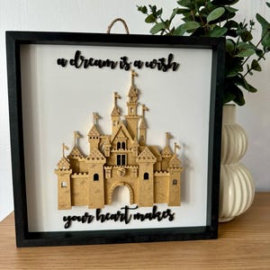 Könnte beinhalten: Eine schwarze gerahmte Wanddekoration mit einem 3D-Papierschnitt von Cinderellas Schloss. Der Text "a dream is a wish your heart makes" ist in schwarzen Buchstaben geschrieben.