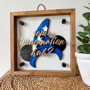 May include: A framed wall art piece with a blue and gold sorcerer's hat and the text "Some imagination huh?" in gold lettering. The hat is layered over a black and white Mickey Mouse silhouette.