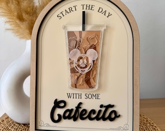 May include: A wooden sign with a Mickey Mouse head on a brown iced coffee cup. The sign reads "Start the day with some Cafecito".