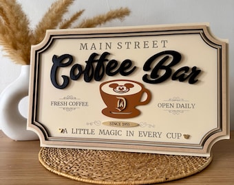 May include: A wooden sign with the text "Main Street Coffee Bar" in black lettering. The sign features a brown coffee cup with Mickey Mouse ears and a gold Disney "D" logo. The sign also includes the text "Fresh Coffee", "Open Daily", "Since 1955", and "A Little Magic in Every Cup".
