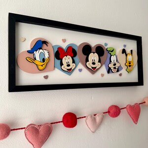 May include: A framed artwork featuring Disney characters Donald Duck, Minnie Mouse, Mickey Mouse, Goofy, and Pluto, each on a heart-shaped background. The characters are in a black frame. A felt heart garland hangs below.