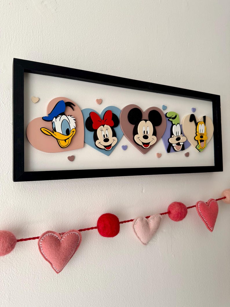 May include: A framed artwork featuring Disney characters Donald Duck, Minnie Mouse, Mickey Mouse, Goofy, and Pluto, each on a heart-shaped background. The characters are in a black frame. A felt heart garland hangs below.