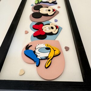 May include: A framed artwork featuring cartoon characters, including Mickey Mouse, Minnie Mouse, and Donald Duck, arranged on heart-shaped backgrounds. The characters are in a 3D style, with a black frame and a white background.
