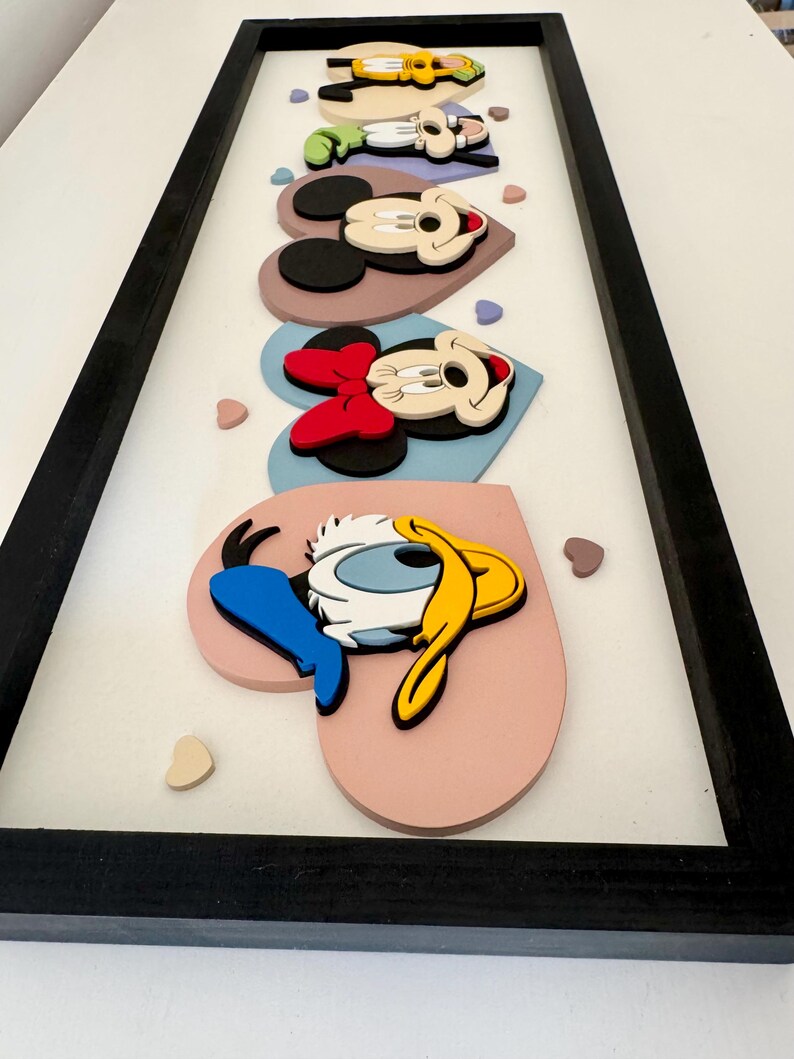 May include: A framed artwork featuring cartoon characters, including Mickey Mouse, Minnie Mouse, and Donald Duck, arranged on heart-shaped backgrounds. The characters are in a 3D style, with a black frame and a white background.