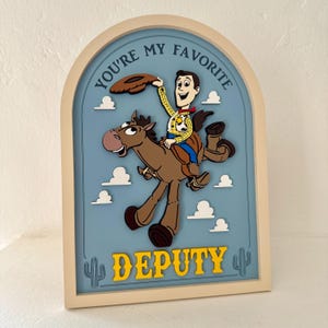 May include: A framed wall art piece featuring a cartoon cowboy riding a horse against a blue sky with clouds. The text "YOU'RE MY FAVORITE DEPUTY" is displayed in yellow. The artwork has a light beige frame.
