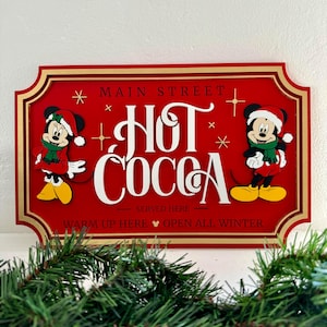 K&ouml;nnte beinhalten: Rotes Holzschild mit goldener Umrandung, auf dem Mickey und Minnie Mouse in Weihnachtsm&uuml;tzen zu sehen sind. Das Schild lautet "Main Street Hot Cocoa Served Here Warm Up Here Open All Winter".