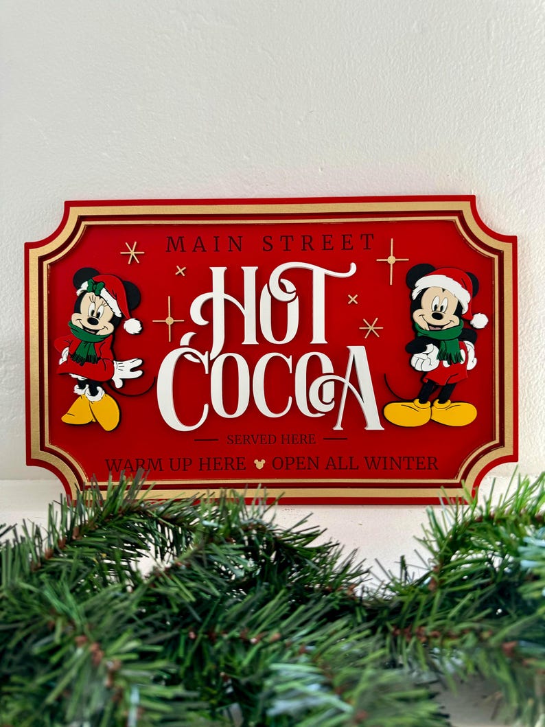K&ouml;nnte beinhalten: Rotes Holzschild mit goldener Umrandung, auf dem Mickey und Minnie Mouse in Weihnachtsm&uuml;tzen zu sehen sind. Das Schild lautet "Main Street Hot Cocoa Served Here Warm Up Here Open All Winter".