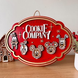 Könnte beinhalten: Ein ovales rotes Holzschild mit Goldrand, das den weißen Schriftzug "MAIN STREET COOKIE COMPANY" und "BAKED WITH MAGIC SINCE 1955" trägt. Es zeigt fünf Lebkuchen-Charaktergesichter, darunter Goofy, Donald, Mickey, Minnie und Pluto.