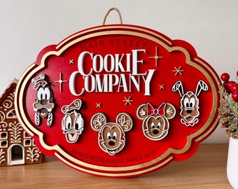 Könnte beinhalten: Ein ovales rotes Holzschild mit Goldrand, das den weißen Schriftzug "MAIN STREET COOKIE COMPANY" und "BAKED WITH MAGIC SINCE 1955" trägt. Es zeigt fünf Lebkuchen-Charaktergesichter, darunter Goofy, Donald, Mickey, Minnie und Pluto.