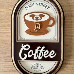 May include: A brown oval-shaped sign with gold trim. The sign says "Main Street Coffee" in a white, cursive font. The sign also features a brown coffee cup with Mickey Mouse's face in the foam and a brown Disney castle logo. The sign says "Magic in Every Cup" in a smaller font.