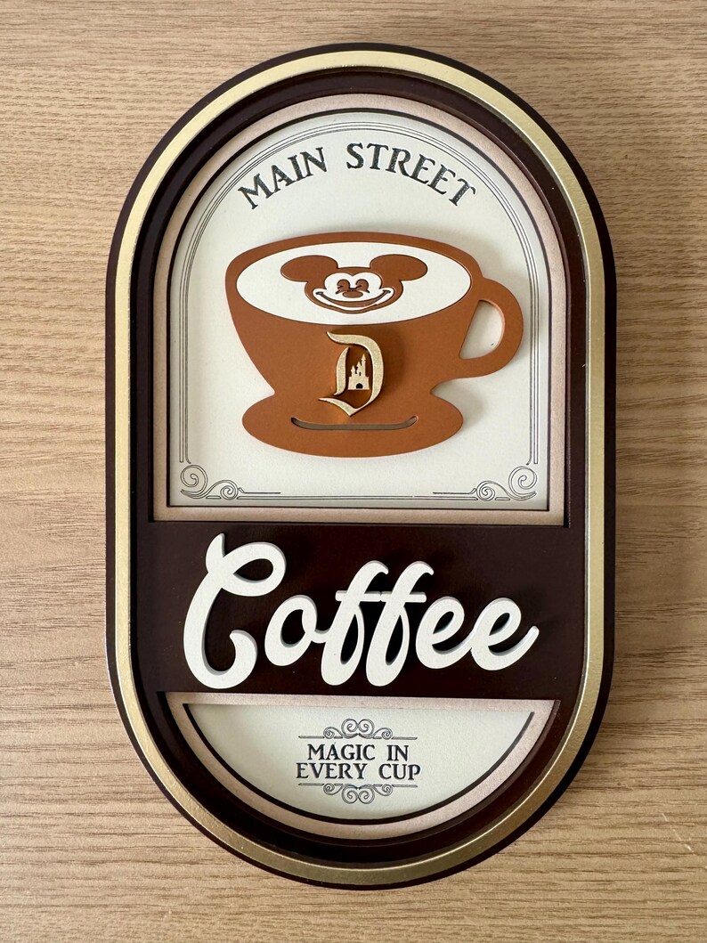 May include: A brown oval-shaped sign with gold trim. The sign says "Main Street Coffee" in a white, cursive font. The sign also features a brown coffee cup with Mickey Mouse's face in the foam and a brown Disney castle logo. The sign says "Magic in Every Cup" in a smaller font.