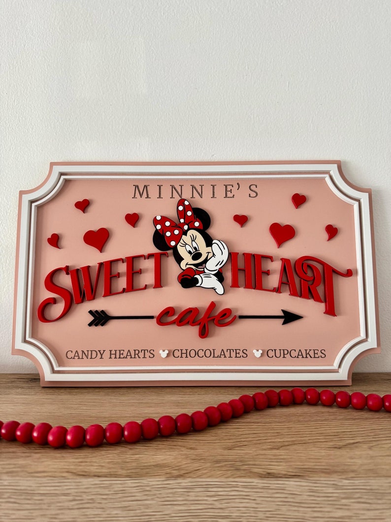 May include: A pink and white sign with a red and white Minnie Mouse illustration. The sign reads "Minnie's Sweetheart cafe" with a red arrow pointing to the right. The text "Candy Hearts Chocolates Cupcakes" is at the bottom of the sign.