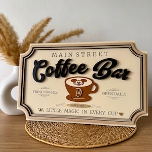 K&ouml;nnte beinhalten: Ein Holzschild mit den Worten "Main Street Coffee Bar" in schwarzer Schrift. Das Schild zeigt eine braune Kaffeetasse mit Mickey-Mouse-Ohren und einem "D" f&uuml;r Disney. Das Schild tr&auml;gt au&szlig;erdem die Aufschrift "Fresh Coffee", "Open Daily" und "Since 1955". Das Schild hat ein goldenes Herz und den Text "A Little Magic in Every Cup".