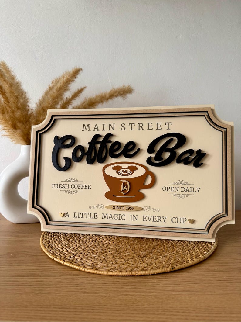 K&ouml;nnte beinhalten: Ein Holzschild mit den Worten "Main Street Coffee Bar" in schwarzer Schrift. Das Schild zeigt eine braune Kaffeetasse mit Mickey-Mouse-Ohren und einem "D" f&uuml;r Disney. Das Schild tr&auml;gt au&szlig;erdem die Aufschrift "Fresh Coffee", "Open Daily" und "Since 1955". Das Schild hat ein goldenes Herz und den Text "A Little Magic in Every Cup".