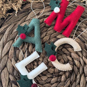 May include: A collection of felt Christmas ornaments spelling out the word "MACS". The letters are in red, green, and white, with holly leaf and berry accents. A red felt heart is also present.