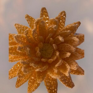 May include: A handmade, crocheted flower in shades of yellow and gold. The flower features multiple layers of petals with a detailed, openwork design. The centre of the flower is a lighter yellow, creating contrast. The overall effect is a textured, three-dimensional floral design.