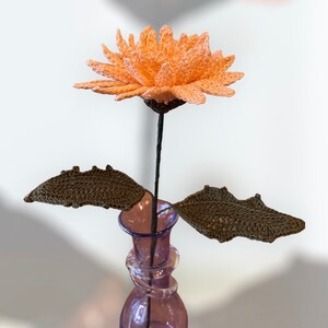 May include: A handmade crochet flower with peach petals, a dark stem, and two brown leaves. The flower is displayed in a purple glass vase. The craftwork is detailed and the flower is a unique decorative item.