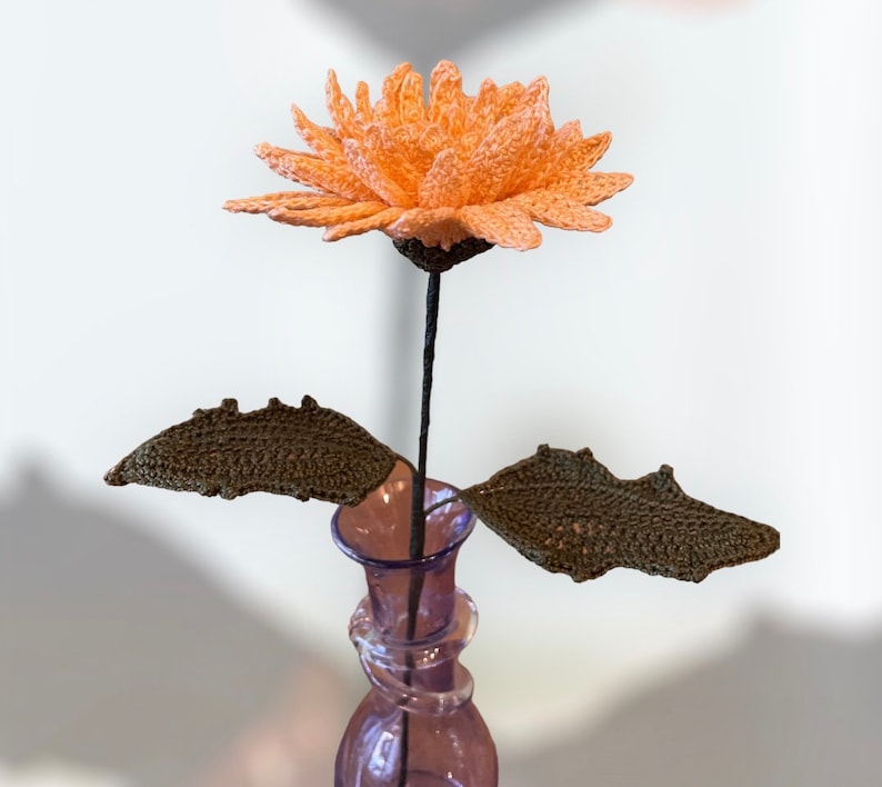 May include: A handmade crochet flower with peach petals, a dark stem, and two brown leaves. The flower is displayed in a purple glass vase. The craftwork is detailed and the flower is a unique decorative item.