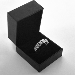 Può includere: Un anello in argento con design a catena, presentato in un cofanetto nero aperto. L'anello presenta maglie a catena interconnesse, creando un motivo continuo. L'interno del cofanetto è scuro.