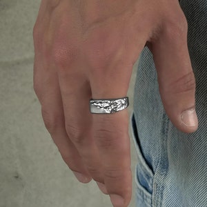 Könnte beinhalten: Ein silberner Ring mit einem strukturierten, organischen Design wird an einer Hand getragen, gepaart mit einem passenden Kettenarmband. Die Oberfläche des Rings hat ein unregelmäßiges, fast geschmolzenes Aussehen. Die Person trägt eine blaue Jeans.