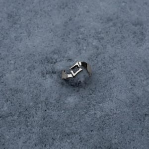 May include: A silver-toned metal ring featuring an open, bypass-style design with two pointed, angular ends that overlap slightly. The band has a smooth, polished finish and a distinct, sharp geometric shape that creates a negative space gap.
