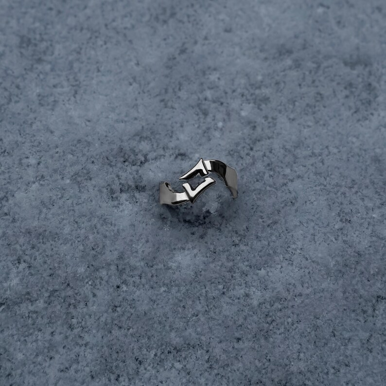 May include: A silver-toned metal ring featuring an open, bypass-style design with two pointed, angular ends that overlap slightly. The band has a smooth, polished finish and a distinct, sharp geometric shape that creates a negative space gap.