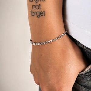 Könnte beinhalten: Ein silberfarbenes Metallkettenarmband bestehend aus einer Reihe miteinander verbundener runder Glieder. Das Armband wird am Handgelenk getragen und zeigt ein schlichtes, gleichmäßiges Design mit einer polierten Oberfläche, die das Licht reflektiert.