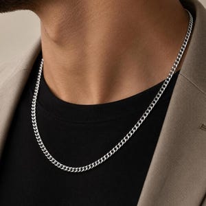 Könnte beinhalten: Eine silberfarbene Panzerkette mit ineinandergreifenden, flachen, ovalen Gliedern. Die metallische Kette liegt auf einem schwarzen T-Shirt und einem beigen Blazer und zeichnet sich durch ein poliertes Finish und ein gleichmäßiges Glieddesign aus.