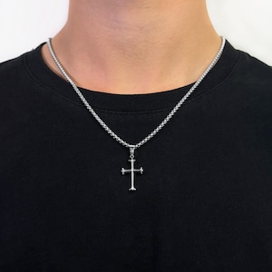 Könnte beinhalten: Eine silberne Kettenkette mit einem silbernen Kreuzanhänger. Das Kreuz hat ein schlichtes Design mit spitzen Enden.