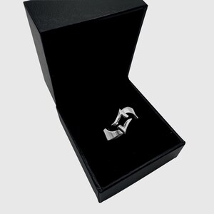 May include: A silver-toned metal ring featuring a stylized, angular design with pointed edges, presented inside an open, rectangular black jewelry box with a dark interior lining. The ring has a smooth, polished finish and a distinct geometric shape.