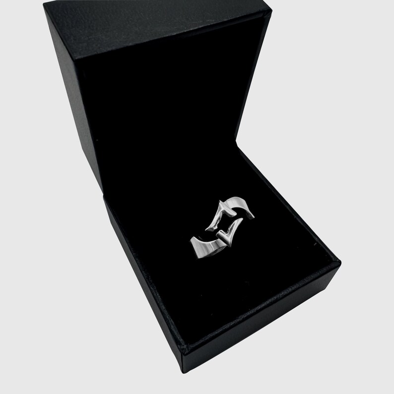 May include: A silver-toned metal ring featuring a stylized, angular design with pointed edges, presented inside an open, rectangular black jewelry box with a dark interior lining. The ring has a smooth, polished finish and a distinct geometric shape.