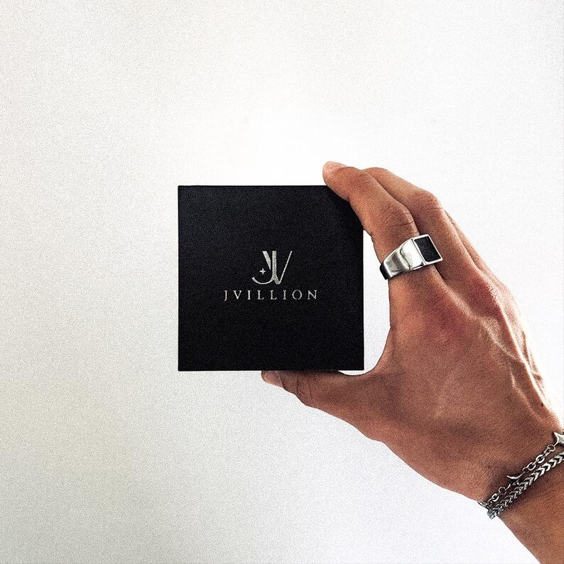 May include: A square black box featuring the silver logo and text JVILLION centered on the front. A silver signet ring with a black rectangular inlay and a silver chain link bracelet are visible on the wrist adjacent to the box.