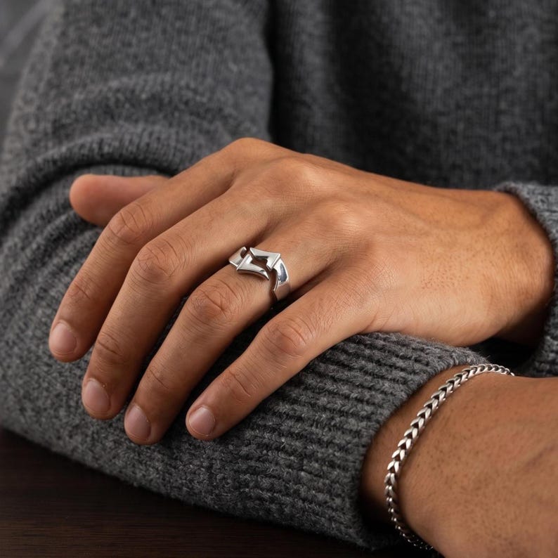 May include: A polished silver ring featuring a geometric, angular design with a central open space, worn on the ring finger. The band has a smooth, reflective metallic finish and a distinct, sharp-edged shape that wraps around the finger.