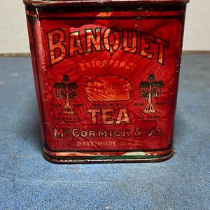 May include: A vintage red metal tea tin with a square shape and rounded corners. The front features the text BANQUET EXTRA FANCY BLENDED GREEN AND BLACK TEA, MCCORMICK & CO. BALTIMORE U.S.A., and two illustrations of hanging lamps.