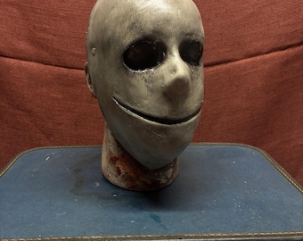 May include: A full-head mask featuring a smooth, pale gray surface with a weathered, textured finish. The mask has large, hollow, dark eye sockets and a thin, horizontal slit for a mouth. The base of the neck area shows dark red, blood-like stains.