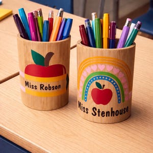 May include: Two wooden pencil pots filled with pens and pencils. One has a red apple design with the text "Miss Robson", the other a rainbow and apple design with the text "Miss Stenhouse". Ideal for a teacher's desk.