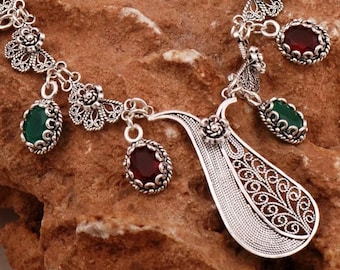 May include: A silver necklace with a large, ornate pendant and multiple smaller pendants with green and red gemstones.