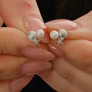 A pair of mini 925 sterling silver stud earrings featuring a detailed seashell and starfish motif accented with a small white pearl and butterfly backs.