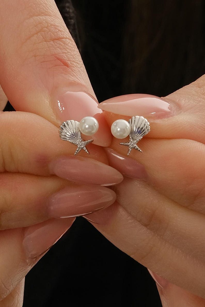 A pair of mini 925 sterling silver stud earrings featuring a detailed seashell and starfish motif accented with a small white pearl and butterfly backs.