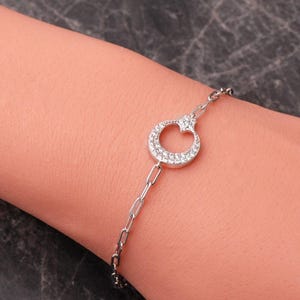 May include: A silver-toned chain bracelet featuring a central circular pendant adorned with small, clear, sparkling stones. The pendant is shaped like a crescent moon with a small star detail at the top, connected to a delicate, elongated link chain.