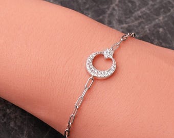 May include: A silver-toned chain bracelet featuring a central circular pendant adorned with small, clear, sparkling stones. The pendant is shaped like a crescent moon with a small star detail at the top, connected to a delicate, elongated link chain.