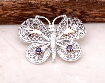 May include: Silver butterfly brooch with filigree wings, gold accents, and two blue gemstone centers. The brooch has a detailed, openwork design and is set against a wooden background.