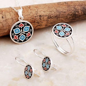 May include: A jewelry set consisting of a circular pendant on a silver chain, a matching ring, and a pair of drop earrings. Each piece features a black enamel background with a symmetrical floral pattern in light blue, red, and white accents.